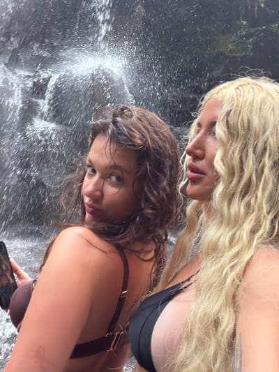 In Gianyar, Indonesia, two individuals are captured in a selfie in front of a waterfall. The person in the foreground, facing the camera, has dark, wavy hair and is wearing a dark-colored bikini top. They are holding a smartphone, suggesting they are taking the photograph. To their right, a second individual with long, blonde, curly hair is visible, also wearing a dark bikini top. The background is dominated by the cascading water and dark rock face of the waterfall, with water droplets creating a misty effect. The lighting appears to be natural, characteristic of an outdoor environment.