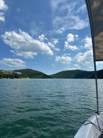 The image depicts a view from a boat on the Abrau Lake in Abrau-Dyurso, Russia. The lake's water is a deep blue-green, with small waves rippling across its surface. In the distance, lush green hills covered in dense vegetation rise from the water's edge. A small settlement with houses and buildings is visible on the slope of one of the hills. The sky above is a vibrant blue, adorned with scattered white cumulus clouds. The right side of the image shows a portion of the boat, including a white hull with purple circular patterns and a metal railing. The overall scene conveys a sense of a peaceful day on the water, surrounded by natural beauty.