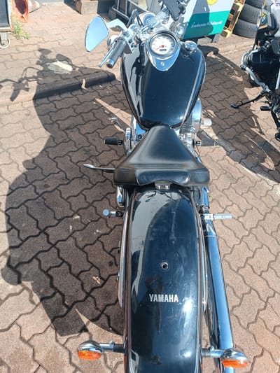 The image shows the rear view of a black Yamaha motorcycle parked on a brick-patterned pavement in Germersheim, Germany. The motorcycle has a prominent chrome exhaust pipe and turn signals. The brand name "YAMAHA" is visible on the rear fender. A shadow of the motorcycle and a person is cast on the ground. In the background, there's a building with a sign that reads "Anerkannter Prüfpunkt" and some wooden pallets with tires. The scene appears to be outdoors, possibly in a parking area or a street.