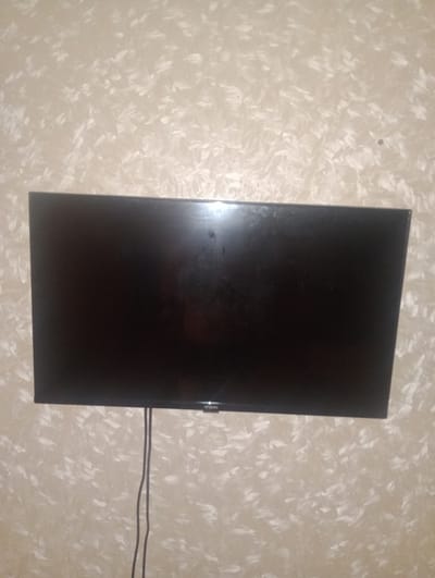 A black flat-screen television is mounted on a textured beige wall. The television is displaying a dark screen, possibly indicating it is turned off or showing a blank image. A black power cable hangs down from the bottom center of the television, descending vertically. The wall has a distinct pattern of brushstrokes or spatters, creating a mottled or abstract effect with lighter and darker beige tones. The lighting suggests an indoor setting, possibly in the evening or with artificial light, as there are reflections on the television screen, indicating light sources in the room. No people are visible in the media. The overall scene is domestic and simple, focusing on the electronic device and its mounting. The location context is Petropavl, Kazakhstan, but no specific visual cues within the image confirm this. No text is visible.