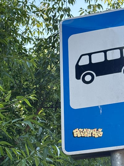 The image shows a street sign with a depiction of a bus and a small, colorful graffiti sticker. The sign is blue and white, and the sticker is yellow with red dots. The sign is partially obscured by green foliage, suggesting it is located outdoors in a natural setting. The sky is visible through the leaves, indicating it is daytime. The graffiti sticker appears to be a stylized representation of text, but it is too blurry to read. The sign is mounted on a metal pole, and the background is a dense arrangement of green leaves and branches. The overall scene appears to be a street in a park or a residential area with trees. There is no indication of any people or specific activity taking place. The weather seems to be fair and sunny.
