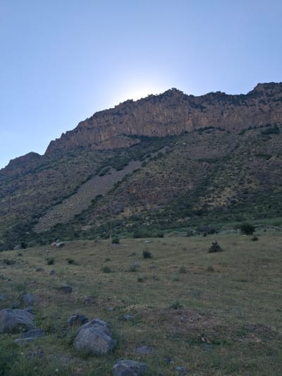 The image depicts a dramatic mountain landscape under a clear, bright sky. The sun is visible, partially obscured by the steep, rocky face of a mountain that dominates the left side of the frame. This mountain is characterized by its rugged texture and layered rock formations. The slope of the mountain is covered in sparse vegetation, with patches of darker green foliage interspersed with lighter, dry grasses and rocks.
To the right of the mountain, a grassy slope descends, dotted with more rocks and a few small, dark shrubs or trees. The grass appears to be a mix of green and dry, suggesting a temperate climate. The overall scene is one of natural wilderness, with no signs of human habitation or activity other than what might be inferred from the terrain itself.
The lighting suggests that it is daytime, likely morning or late afternoon, given the angle of the sun. The weather appears to be clear and fair. The location context provided, Shatin, Armenia, places this mountainous region in the Armenian Highlands, known for its dramatic topography. There is no visible text in the image. The composition, with the mountain rising steeply against the expansive sky, evokes a sense of awe and the grandeur of nature.