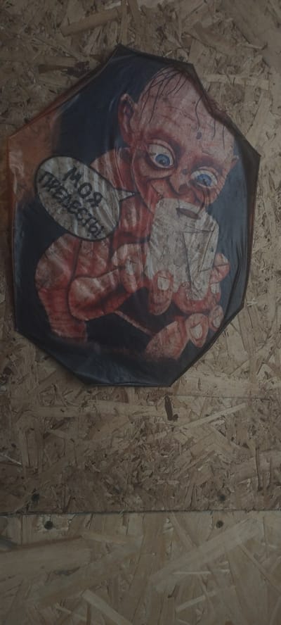 The image shows a printed umbrella hanging on a wooden wall. The umbrella features a stylized drawing of the character Gollum from "The Lord of the Rings," with blue eyes and a speech bubble containing the Russian text "Моя прелесть!" which translates to "My precious!". The wall appears to be made of oriented strand board (OSB) and is likely located indoors, as indicated by the flat, consistent lighting. The specific location in Покосное, Russia cannot be determined from the image alone.