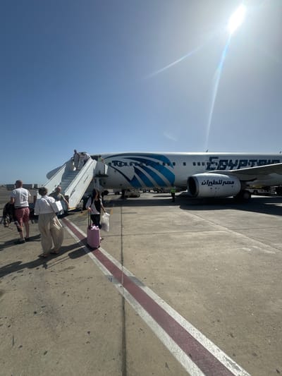 The image depicts an EgyptAir airplane on a concrete tarmac under a clear blue sky, with a bright sun in the upper left corner. Passengers are disembarking via a set of stairs connected to the aircraft. Several people are walking down the stairs, while others are on the ground, with luggage including a pink suitcase. The side of the plane shows the EgyptAir logo in both English and Arabic. A single person in a high visibility vest is standing near the plane. A red and white striped line is visible on the tarmac. The location is the city of Unknown, Egypt.