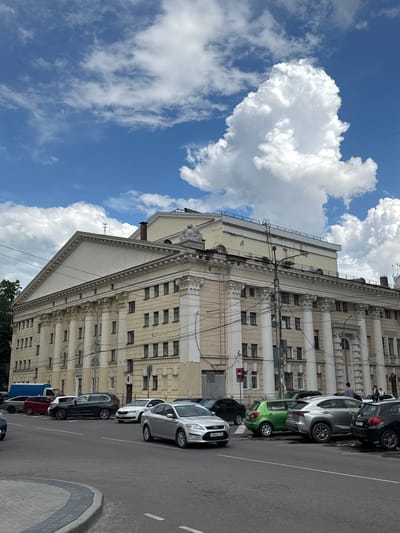 The image depicts a street scene in the посёлок 1-го отделения совхоза "Масловский", Russia. A large, ornate building with classical architectural elements, characterized by columns and numerous windows, dominates the left side of the frame. The building appears to be older, with some signs of wear and tear visible on its facade. A bright blue sky with large, fluffy white clouds fills the upper portion of the image.
In the foreground and along the right side of the street, several cars are parked in a row. These include a silver hatchback, a green hatchback, a dark sedan, a red sedan, and a white sedan with visible license plates. A traffic light with a red signal is also visible near the parked cars. Overhead wires are strung across the scene, connecting to the building and a utility pole. The presence of multiple cars and the street infrastructure suggest this is an urban or semi-urban setting.
The weather appears to be sunny and clear, with bright daylight casting shadows. The clouds suggest a pleasant, possibly warm, day. The overall mood is neutral and observational, capturing a typical street view. There is a license plate visible on one of the cars that reads "Y680BY 126".