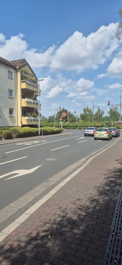 The image depicts a street scene in Mainz, Germany. The street is asphalt with white lane markings and a pedestrian walkway paved with brick. A light yellow apartment building with multiple balconies filled with plants stands to the left of the road. A street lamp is positioned in front of the building. Several cars are visible on the road, including a silver sedan, a light green hatchback, and a dark sedan. Traffic lights and a stop sign are present at an intersection ahead. The sky is blue with scattered white clouds, and trees and green bushes line the side of the road. The overall impression is a sunny day in a residential area.