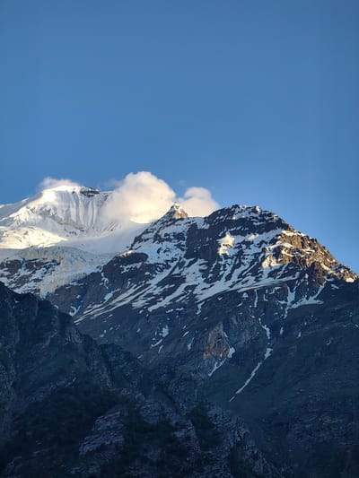 This image captures a majestic mountain range under a clear, vibrant blue sky. The peaks are partially covered in snow, with dramatic shadows and highlights indicating the presence of sunlight. The time of day appears to be either early morning or late afternoon, given the angled light casting long shadows and illuminating the snow-capped summits.
The foreground is dominated by dark, rugged rocky slopes, some with patches of green vegetation hinting at lower elevations. As the eye moves upward, the terrain transitions to rocky outcrops and scree slopes, increasingly covered by snow. The middle ground features a large, imposing mountain peak with distinct snowfields and crevasses, suggesting a glacier or significant snow accumulation. Further back, another, larger peak rises, its upper reaches completely covered in white snow and touched by the sun, creating a brilliant contrast against the sky. A few wispy clouds drift near the summit of this larger peak, adding a sense of depth and atmosphere.
There are no people or specific objects visible in the frame, focusing the attention entirely on the natural grandeur of the landscape. The overall mood conveyed is one of awe and tranquility, highlighting the immense scale and beauty of the natural world. The location is identified as Kead, India. The weather is clear, with bright sunshine illuminating the mountains. No text is visible in the image.