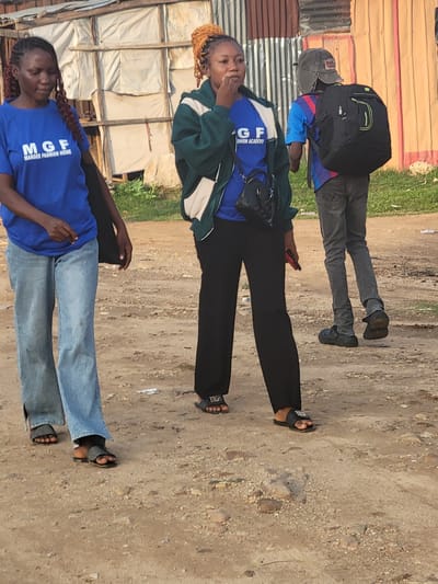 Three people are standing outdoors on a dirt surface that is likely a road. Two of the individuals are wearing matching blue t-shirts with the text "MGF MAROBE FASHION HOME" printed on them. The first person is wearing blue jeans and the second person is wearing black pants. The third person is wearing a blue jacket, grey jeans, and a black backpack. The buildings in the background are of simple construction, possibly made from metal sheets and timber. The image appears to be taken during the day with natural light. The location is Kwamba, Nigeria.