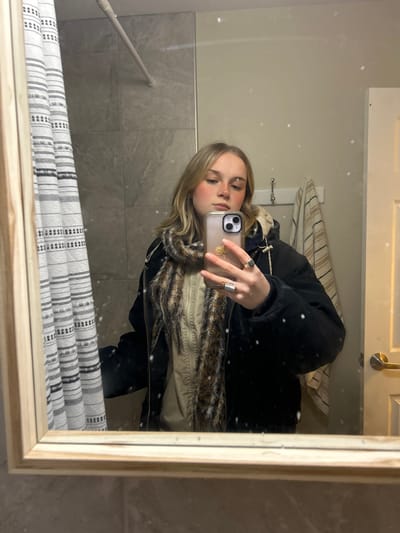 A mirrored selfie shows a young woman wearing a black jacket with a fur-lined collar. She holds a phone with a floral-patterned case. The background features a tiled wall and a striped shower curtain. A towel rack and a cabinet are also visible in the reflection. The image is slightly rotated and has numerous white, sparkling dots overlaid. The frame of the mirror is a light wooden color.