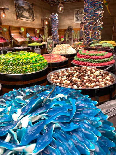 This image depicts a vibrant candy store, likely located in Dubrovnik, Croatia, given the presence of an advertisement for "Captain Candy" which is a known candy shop in Dubrovnik. In the foreground, a large pile of blue and white dolphin-shaped gummy candies is displayed, with a silver candy tong resting on top. Behind this, several barrels are filled with various candies. To the left, a barrel overflows with bright green and yellow gummy candies. To the right of that, a barrel is filled with a mix of chocolate and white spherical candies. Further back, more barrels showcase colorful candies arranged in circular patterns, including red and green layered candies resembling slices of fruit, and mounds of white and pink confectioneries. In the background, a wooden wall displays framed artwork and other advertisements. The lighting is warm and inviting, typical of a retail environment. Two individuals are visible in the background, partially obscured by the candy displays. One appears to be a woman with long brown hair, and the other is a man. The overall impression is one of abundance and variety within a themed candy shop.