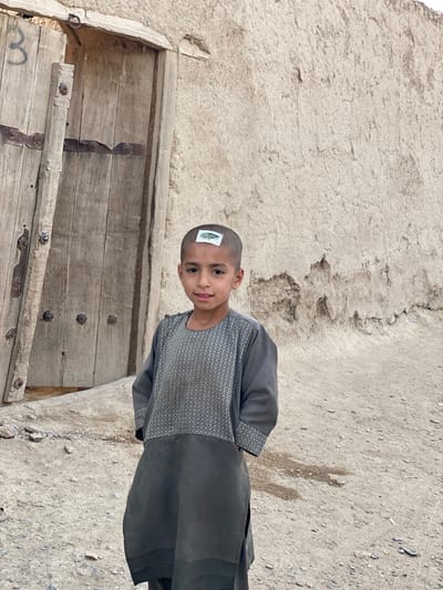 A young boy with a shaved head stands in front of a weathered wooden door. He is wearing a dark gray tunic with a patterned bib. A small sticker, possibly a temporary tattoo, is affixed to the top of his head. The boy is looking at the camera with a slight smile. The background consists of a rough, textured wall made of mud or plaster, and the ground is covered in dirt and small stones. The scene appears to be outdoors, likely in a village setting, given the rustic architecture and the arid ground. The lighting suggests it is daytime, possibly a sunny day due to the shadows, although the overall mood is muted. The image captures a candid moment of the boy. There is no visible text in the image.
