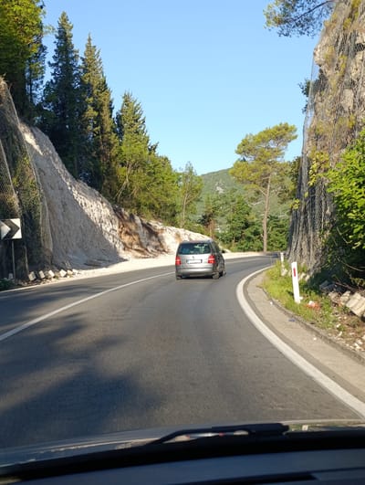 The image depicts a view from inside a vehicle looking out at a winding road on a sunny day, likely in the vicinity of Kruševica, Montenegro. The road curves to the right, with a gray car in front. On the left side of the road, a steep, light-colored embankment is reinforced with wire mesh, topped by coniferous trees. On the right side, another rocky embankment, also secured with mesh, is covered in dense green foliage. The sky is clear and blue. Several white road markers with red tops are visible along the edge of the road. The foreground shows the dashboard and windshield wipers of the vehicle.