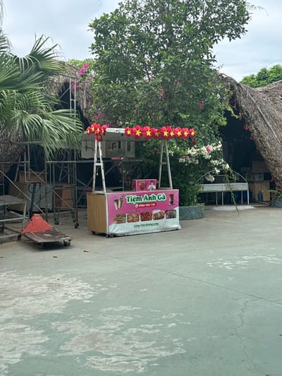The image depicts an outdoor scene in Hoa Lư Ward, Vietnam. In the foreground, a pink and white food cart is set up, displaying various food items and beverages. The cart has signage with Vietnamese text and images of drinks and food. Above the cart, a string of red and yellow star decorations hangs, possibly indicating a festive occasion. Behind the cart, a thatched-roof structure is visible, suggesting a rustic or traditional architectural style. To the left, a red hand truck and a metal trolley are present, indicating potential loading or unloading activities. Lush green trees and foliage surround the area, with blooming pink and white flowers adding color to the scene. The ground is paved with concrete, showing signs of wear and tear. The overall impression is that of a casual outdoor food vendor operating in a tropical or subtropical environment.