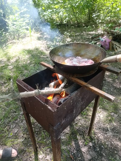 In Shumeika, Russia, a rustic outdoor cooking scene unfolds. A weathered metal grill, possibly homemade, contains a lively fire with glowing embers and dancing flames, topped by logs and branches. Suspended over the fire on metal supports is a large wok-like pan filled with raw, pink chunks of meat, shimmering in the heat. Wisps of smoke curl upwards from the pan, mingling with the dappled sunlight filtering through the lush green foliage of trees that surround the area. The ground is covered in a mix of dry leaves, twigs, and patches of grass, indicating a natural, possibly wooded setting. In the lower left corner, a person's foot clad in a black flip-flop is visible, suggesting a casual, informal gathering. The overall atmosphere is one of a simple, outdoor meal preparation, likely during the daytime with fair weather allowing for this al fresco activity. No text is visible in the image.