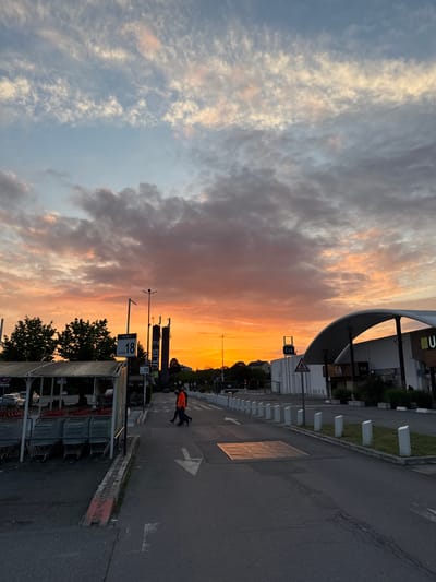 The image is taken in Les Ulis, France, at sunset. The view is from a parking area beside a shopping center. To the right is an aisle of shopping carts. To the left is the road to the parking area, where there is a person in an orange vest walking. Beyond the road, there is a building that features "CA" on its signage. The sky is filled with clouds lit by the setting sun.