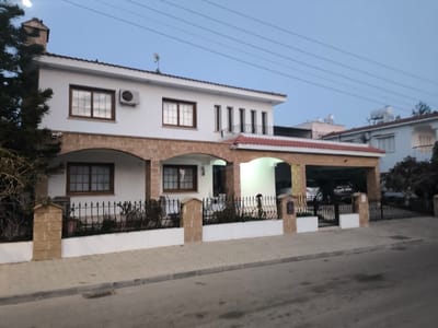 A two-story house with a white facade and brown-framed windows stands behind a low fence. The house features arched doorways and a carport. A chimney is visible on the left side of the roof. The sky is overcast, and power lines are stretched across the scene. A paved street is in the foreground. Other houses are visible in the background.