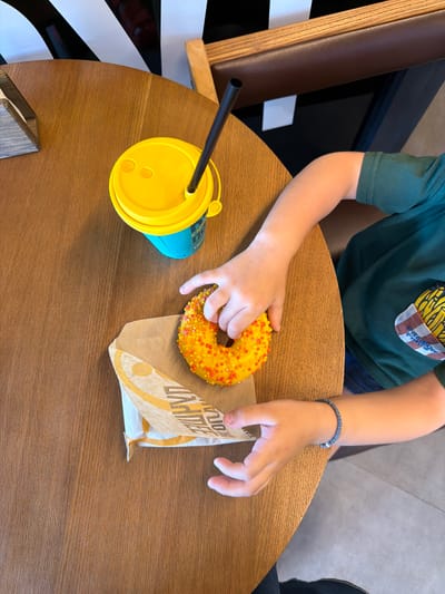 An overhead shot shows a child's hands on a wooden table. The child is wearing a green t-shirt and a beaded bracelet. In front of the child are a yellow donut with orange and yellow sprinkles, a brown paper wrapper, and a blue and yellow cup with a black straw. The scene appears to be in a cafe or restaurant, possibly in Tikhoretsk, Russia.