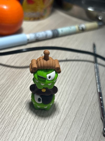 A small, green, cartoonish figurine is the primary subject of this image. It appears to be a character with large white eyes outlined in black, a small black nose, and white dots scattered across its body. The figurine has brown, segmented appendages, one of which extends from its front like a handle. Behind the figurine, a black cable snakes across a light-colored, wood-grained surface. To the left of the figurine, a long, slender object with a light blue casing and some blurry text is visible. Further to the left, a blurry orange object and part of a yellow package with red and green elements can be seen. At the top of the image, a silver metal object with a ridged surface is present. The scene appears to be indoors, with the lighting suggesting it could be daytime. No specific location cues are visible beyond the general indoor setting and the wood-grained surface. The figurine's expression is neutral or perhaps slightly surprised due to its wide eyes. There is no clear activity or event taking place; the objects appear to be resting on a surface.