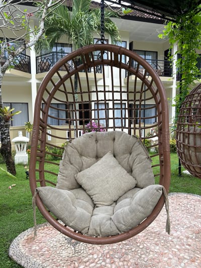 The image depicts a wicker hanging chair with thick, light-colored cushions and a square pillow, positioned outdoors on a circular mosaic of small, polished stones. Behind the chair, a manicured lawn is visible, with a white stone lantern on the left and a cluster of pink flowers and green foliage to the right. In the background, a multi-story building with light-colored walls, balconies with black railings, and tiled roofs is visible. Palm trees and other greenery surround the building and the immediate outdoor area. The scene suggests a resort or hotel setting in Karangasem, Indonesia, designed for relaxation in a tropical environment. There are no people present in the image.