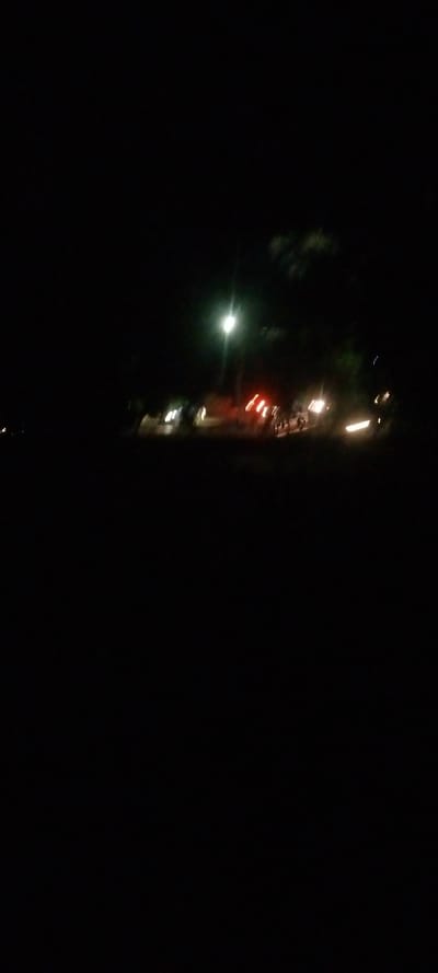A nighttime street scene shows blurry vehicles and lights scattered along a road. A central streetlight is visible, casting a glow amidst the darkness. Red taillights suggest braking cars, while other white lights indicate headlights. Silhouetted trees are barely distinguishable against the night sky. The foreground is dominated by darkness.