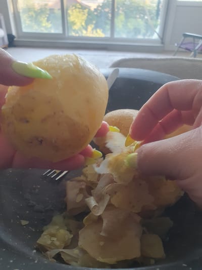 In a close-up shot, a person's hands with bright neon green painted nails are peeling a cooked potato over a dark bowl filled with peeled potatoes and their skins. The potato being held is round and pale yellow, with a few brown speckles. The other hand is actively peeling another potato, with pieces of skin curling away. The bowl appears to be made of speckled dark material and contains several chunks of cooked potato and loose skins. A fork with metal tines is partially visible in the lower left corner of the bowl. In the background, a large window reveals a blurry scene of green trees and buildings under a bright sky, suggesting an indoor setting with natural light. The overall impression is of food preparation. The location is not discernible from the image.