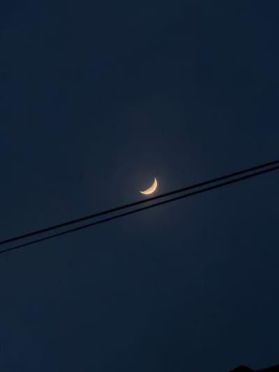A dark blue night sky holds a crescent moon. The moon is small and brightly lit, appearing crescent-shaped. Two black power lines run diagonally across the frame, partially obscuring the moon. The lighting is dim, and the overall tone of the image is tranquil and subdued.