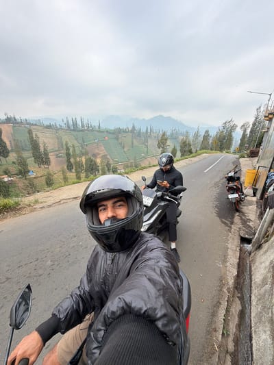 The image shows two individuals on motorcycles on a paved road in Ngadas, Indonesia. The person in the foreground is taking a selfie, wearing a black helmet and jacket, with a landscape of terraced fields and trees visible behind them. The second individual, further down the road, is also on a motorcycle, wearing a black hoodie and helmet, and is looking at their phone. The road curves to the right, and on the right side of the road, there is a concrete wall with some items piled against it, including a yellow bin and what appears to be fabric. The sky is overcast, and the overall environment suggests a rural mountainous region.