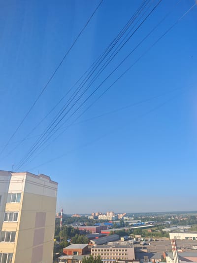 An aerial view captures the city of Voskresenski, Russia, on a clear day. The foreground features a portion of a tall, light-colored apartment building with beige and pale pink panels. Beyond the building, the cityscape unfolds with a variety of structures including industrial buildings with red roofs, smaller residential buildings, and a prominent tall chimney with red and white stripes. Dense green trees are interspersed throughout the urban landscape. Overhead, a series of thin, dark power lines crisscross the bright blue sky, creating a distinct visual element. The horizon is flat, suggesting a generally level terrain. The image appears to have been taken from a high vantage point, possibly another building.