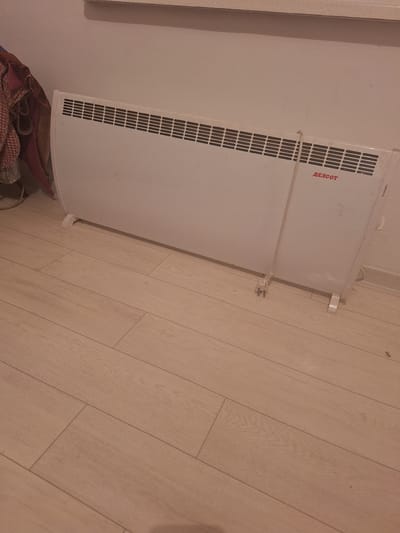 A white electric heater is positioned on a light-colored wooden floor against a pale wall. The heater has a slatted grille across its front and a small red logo that reads "ДЕЛСОТ" on the right side. A white power cord is plugged into the heater and extends downwards, ending in a plug. To the left of the heater, a partially visible pile of fabric, possibly clothing or blankets, is draped over a surface. The environment appears to be indoors, in a room in a new building in Novostroyka, Kyrgyzstan. The lighting is dim.
