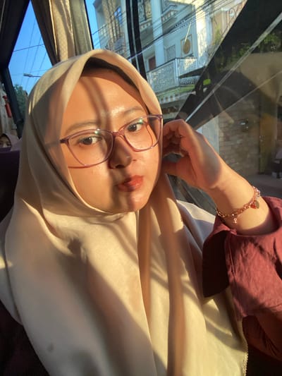 The image captures a person, likely female, from the chest up, positioned inside a vehicle, likely in the city of Batu, Indonesia. The central subject wears a light beige hijab, pink-rimmed glasses, and a maroon long-sleeved garment. Her lips are painted a reddish-orange, and a small nose piercing is visible on her left nostril. Her right hand is raised, with fingers lightly touching her ear/jawline, and a multi-strand, rose gold-colored bracelet with charms adorns her right wrist. Strong, diagonal sunlight illuminates the right side of her face, hijab, and arm, casting distinct shadows. Her gaze is directed slightly to the viewer's right. In the lower-left background, part of another individual, also appearing to wear a hijab, is visible. The vehicle's interior shows a golden-beige curtain in the upper-left. Through the vehicle's window, an urban landscape is visible under a clear blue sky. Multi-story buildings with white facades, decorative pillars, and a balcony with ornate railings are seen on the right, interspersed with green foliage. Overhead electrical wires crisscross the sky. Strong reflections and glare are visible on the window glass, consistent with direct sunlight.