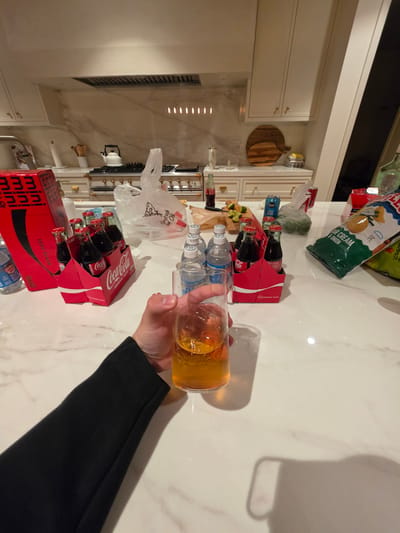 The image displays an indoor kitchen scene, primarily focused on a white marble countertop filled with an assortment of beverages and snacks. In the foreground, a person's right hand, wearing a black long-sleeved garment, holds a clear glass bottle containing a golden-amber liquid.

The countertop features two 6-packs of Coca-Cola Original Taste in glass bottles, and a 6-pack of Coca-Cola Zero Sugar mini cans, with visible text "mini mini mini" and "ZERO SUGAR." Several plastic water bottles, some labeled "PURE CLEAN," are also present. Snacks include a large bag of Lay's potato chips, clearly marked "PURE CREAM & ONION FLAVORED," and another partially visible green bag of chips. A white plastic bag from "Kentucky Fried Chicken" (KFC) is seen, along with a bottle of "CANAD DRY" soda and some limes on a wooden cutting board.

In the background, white kitchen cabinets with gold handles line the walls, and a stove with a white kettle is visible. Two decorative wooden cutting boards hang on the wall. The scene suggests a casual gathering or meal preparation, with a variety of drinks and snacks available. The lighting indicates an indoor setting, likely under artificial light. No specific location cues for Coronado are visible within the image itself.