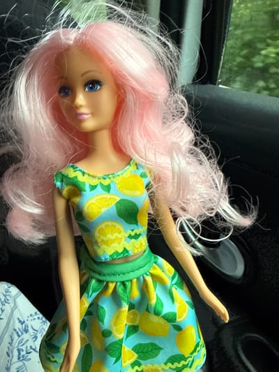 A Barbie doll with long, wavy pink hair is shown from the chest up, sitting in the passenger seat of a car. The doll is wearing a two-piece outfit: a short-sleeved crop top and a skirt, both with a blue background and a pattern of yellow lemons and green leaves. The doll's head is turned to its left, and its eyes are open and looking forward. The background of the image shows the interior of the car, including a dark gray seat, a car door, and a window with a blurry green view of trees outside. The doll appears to be sitting near the center of the frame, with its body slightly angled. The lighting suggests it is daytime, possibly with sunlight filtering through the car window. The location in the city of Weidenhausen, Germany is not discernible from the image itself.