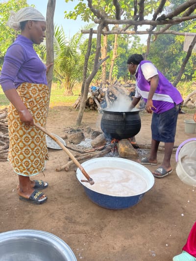 The image depicts an outdoor cooking scene in Tabu, Nigeria, with two individuals engaged in preparing food or drink using large pots over open fires on a bright day.
In the foreground, a woman, wearing a purple long-sleeved top, a patterned yellow and brown skirt, and a white headscarf, is stirring a thick, creamy, foamy liquid in a large blue and white pot with a long wooden stick. Her feet are visible, wearing black slides with "adidas" text.
Adjacent to her, another person, dressed in a purple and white short-sleeved top and dark shorts, bends over a large black pot set directly over a wood fire. Steam rises from this pot, indicating active cooking. This individual also wears black Adidas slides. Piles of firewood are stacked nearby, serving as fuel for the fire.
The setting is an unpaved, sandy ground, surrounded by lush green trees and foliage under a clear sky, characteristic of a rural or village environment. Simple wooden posts and structures are also visible, possibly part of a makeshift shelter or cooking area. Various plastic buckets and basins are scattered around the workspace. The scene portrays a method of communal or large-scale cooking prevalent in the region.
