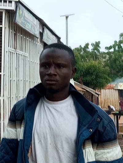 The image is a medium shot of a Black man with short, dark hair and a goatee, wearing a white t-shirt and a blue and white striped jacket. He is looking directly at the camera with a neutral expression. The man is standing in front of a white metal gate, which appears to be part of a shop. Behind the gate, there is a building with a striped awning and some lettering that is partially obscured. In the background, there are trees, a lamppost, and other buildings, suggesting an outdoor setting in a city or town. The overall impression is of a candid street portrait.