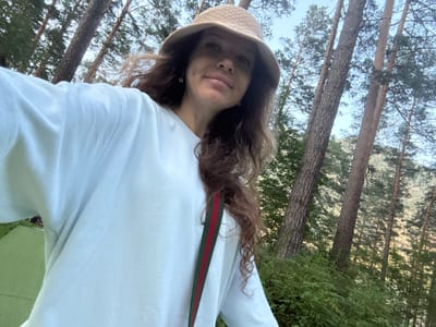 A woman is taking a selfie outdoors in a forest. She is wearing a light blue long-sleeved shirt and a straw bucket hat. A green and red striped strap is visible across her shirt. She has long, wavy brown hair. The background shows tall pine trees with green foliage and a bright sky. There is also a patch of green grass visible on the left side of the image. The scene appears to be located in the city of Belokurikha, Altai Krai, Russia.