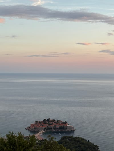 This media file depicts a scenic view of a small island settlement with red-tiled roofs, connected to the mainland by a causeway. The island is nestled in the sea, with a lush green hillside on the right side of the frame. The water is a calm, muted blue-grey, with subtle ripple lines suggesting a gentle breeze. The sky is a soft gradient of pale blue and pastel pinks, indicating either sunrise or sunset. Scattered clouds, tinged with darker grey and hints of orange, drift across the sky. The overall mood is serene and tranquil. The architecture of the settlement is traditional, with stone buildings clustered together on the rocky island. A few small boats are visible in the water near the island. The image captures a moment of natural beauty and architectural charm. The time of day appears to be either dawn or dusk, given the soft lighting and pastel colors in the sky. The weather seems mild and pleasant. The location is identified as Čelobrdo, Montenegro. There is no visible text in the image.