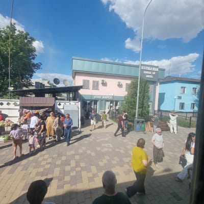 The image depicts a street scene in Turkistan, Kazakhstan, on a sunny day. A light blue building with white window frames is visible on the right, with a sign that appears to read "Nurly-Jol" and "Hotel". To its left is a pastel pink building with a green trim, labeled "KIPI MARKET". In front of these buildings, a paved area is populated with people. Several individuals are browsing goods displayed at a market stall on the left, where fruits and watermelons are visible. Other people are walking on the pavement or standing in groups. The sky is blue with scattered white clouds. The scene appears to be an outdoor marketplace and a commercial area in the city.