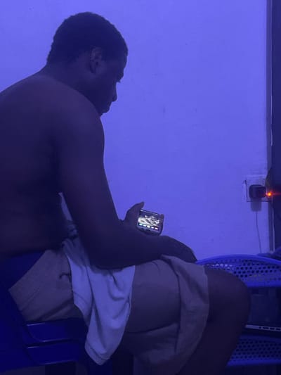 A shirtless individual is reclined in a blue chair, their legs crossed. They hold a phone in their hand, the screen illuminated with what appears to be an image of a lizard. A white t-shirt is draped over their lap, partially obscuring light-colored shorts or underwear. The scene is bathed in a vibrant blue light, and a wall outlet with a red light is visible in the background. A blue basket is also visible.