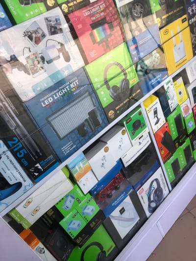 This image captures a vibrant display case filled with numerous packaged electronic gadgets and accessories, likely in a store or market in Epe, Nigeria. The products are neatly arranged in their retail boxes, creating a mosaic of colors and brands. The surface of the display case is reflective, showing partial reflections of surrounding objects, including what appears to be a car wheel.
The variety of items on display includes:
- **Headphones and Audio Devices:** Boxes for JBL Tune M16BT headphones, Oraimo "Endless Beats, All Day Long" headphones, "rexi BLUETOOTH EARPHONES," and AirPods Pro.
- **Gaming Accessories:** Several Sony Dualshock 3 and Dualshock 4 wireless controllers for PlayStation consoles (PS3, PS4), alongside generic wireless controllers.
- **Power and Charging Solutions:** Oraimo power banks with capacities like 20000mAh and 33000mAh, various 6A fast chargers, "Xcharge" fast chargers, Infinix PD Fast Chargers, and Oraimo RK-020 adapters.
- **Photography and Videography Gear:** "PROFESSIONAL PHOTO & VIDEO LED LIGHT KIT" boxes (Model LED-416, with adjustable brightness and color between 3200-5600K), "VIDEO-MAKING KIT" boxes, "SELFIE STICK" packages, and a stand capable of 215 cm extension with "ONE CLICK POP-UP STABLE PAN TILT."
- **Other Electronics:** A yellow box for "MTN Broadband," a black "24G" wireless mouse, EarPods, and "THE PHONE HOLDER."
Visible text includes product names, brands such as SONY, JBL, Oraimo, Infinix, rexi, MTN, and Xcharge, along with specifications like "365 DAYS," "Adjustable Brightness & Color," and "STABLE CURRET." The overall scene suggests a retail environment specializing in consumer electronics.