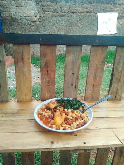 A plate of food sits on a wooden table. The food consists of corn, beans, and a potato, all in a tomato-based sauce. A fork is sitting on the plate. There is a small amount of green leafy vegetables on top of the food. The table is made of wood and is propped up by vertical wooden slats. There is a wall behind the table, with a white piece of paper hanging on the wall. Green grass is visible in the background. This scene is most likely located in a home or a restaurant in Rungiri, Kenya, based on the provided coordinates (-1.248, 36.683).