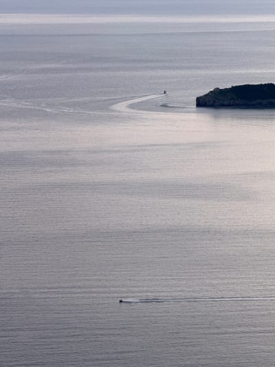 An aerial perspective captures the serene waters surrounding Čelobrdo, Montenegro. The image showcases two small boats navigating the calm sea. One boat, positioned in the lower third of the frame, leaves a subtle white wake as it travels horizontally. Further in the distance, a second boat creates a more pronounced, curving wake as it moves towards a small, rocky island dotted with vegetation. The island itself is a dark mass against the lighter expanse of water, with a small structure visible on its summit. The water's surface is textured with gentle ripples, reflecting the muted light of the sky, which appears overcast and hazy, creating a soft, diffused illumination across the scene. The overall atmosphere is one of tranquility and vastness.