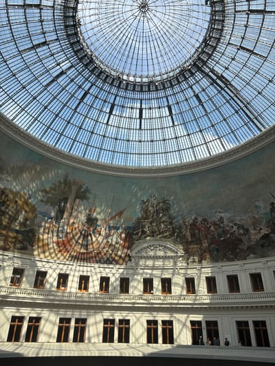The image depicts the interior of the Bourse de Commerce in Paris, France, specifically focusing on its grand glass dome and the surrounding mural. The dome, a marvel of 19th-century engineering, is a latticework of metal and glass, through which a clear blue sky is visible. Sunlight streams through the dome, casting intricate shadows on the curved wall below, creating a dappled effect across a large, colorful mural.
The mural itself is an allegorical depiction of commerce and trade, featuring numerous figures engaged in various activities. Scenes of historical significance, including maritime trade and agricultural labor, are rendered in a classical style. The mural is divided into sections, with a central sculptural element beneath a prominent arch. The colors are rich and varied, though softened by the lighting and shadows.
Below the mural, the building's architecture is visible, characterized by multiple levels of white walls and rows of windows. The windows, framed in wood, are mostly dark, suggesting they lead to interior spaces. A balcony or walkway runs across one level, with decorative balustrades. In the lower portion of the image, several people are visible standing on a walkway, looking towards the center of the building. They are casually dressed, indicating they are likely visitors or patrons. The overall atmosphere is one of grandeur and historical preservation.