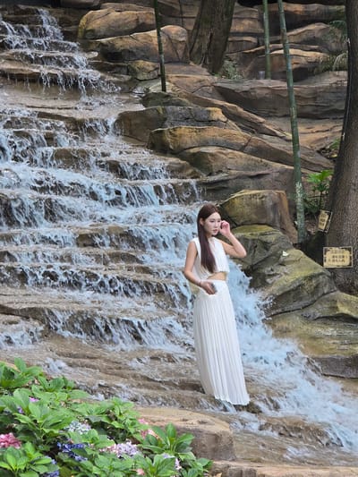 A woman in a white dress stands on rocks beside a tiered waterfall. She is looking to her left, with her right hand near her face. The waterfall cascades down rocks, and green plants, some with pink and purple flowers, are in the foreground. Bamboo stalks are visible behind the woman, and two signs with Chinese characters are posted on the rocks to the right. The scene appears to be in a park or garden setting within the Xiaoshan District of China.