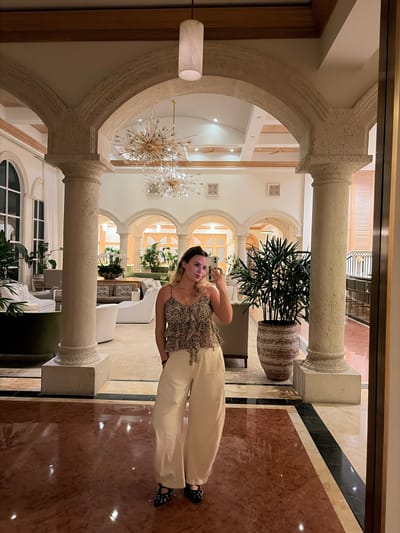 A woman is taking a selfie in an opulent lobby. She wears a patterned top and light-colored pants. The lobby features stone arches supported by pillars, ornate chandeliers, potted plants, and marble flooring. The location appears to be in Key Biscayne, United States of America.