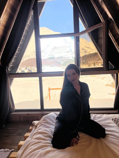 The image displays an indoor scene within an A-frame structure, likely a cabin or chalet, situated in the mountainous region of Virak, Montenegro. A young adult female with long, dark brown hair, wearing black pajamas with white piping, kneels on a bed in the foreground. She is positioned slightly to her left, facing the camera with a subtle smile. The bed is covered with a thick, light-colored duvet and features a visible wooden pallet-style frame.

Behind the subject, a large A-frame window, framed in dark brown, reveals a panoramic view of the exterior. One pane in the upper right section of the window is open, angled inwards. Dark brown curtains are partially drawn on either side of the window. The interior walls and slanted ceiling consist of light-colored wood paneling. A wooden floor and a small, light-colored patterned rug are visible on the left side of the frame.

The outdoor environment visible through the window is a sunny, snowy landscape beneath a clear blue sky. Distant mountains, partially snow-covered with areas of exposed rock and sparse brown vegetation, dominate the background. In the middle distance, several small, dark-roofed A-frame cabins are visible on the left, nestled in the snow. A wooden swing structure is also discernible on the snowy ground further right. Directly outside the window, below the landscape, a dark, corrugated roof is visible.