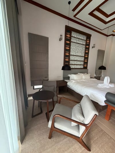 This is a forensic, matter-of-fact description of a hotel room in Ban Kao Dua, Thailand. The room is furnished with a bed, two nightstands, a chair, and a small table. The bed is covered with white linens and has two pillows. A folded towel in the shape of a swan is placed on the right side of the bed. Two black pendant lights hang from the ceiling above the bed. A tall, full-length mirror is positioned to the right of the bed. A large, grey Roman blind covers a window to the left of the bed. A grey armchair with a wooden frame and a light grey cushion sits in the foreground, to the left of the bed. A small, dark brown, round wooden table with a dark brown base stands in front of the armchair. A grey upholstered panel is mounted on the wall behind the armchair. The walls are painted white, and the ceiling features decorative wooden beams arranged in a geometric pattern. A patterned fabric panel is mounted on the wall above the headboard of the bed. A tall, thin object, possibly a floor lamp or a decorative piece, is visible in the far right corner of the room. The flooring appears to be a textured, light brown carpet.