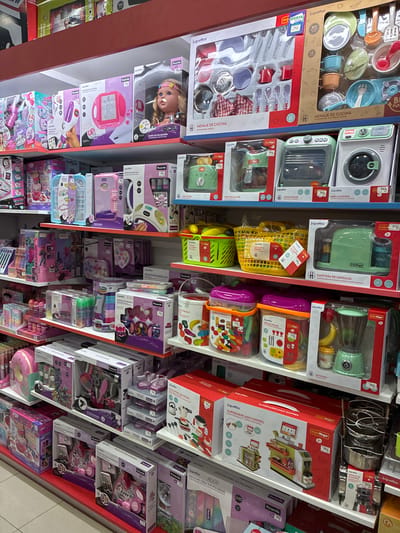 The image displays a well-lit retail store aisle, likely a toy store, in L'Hospitalet de Llobregat, Spain. The shelves are packed with colorful toy sets, predominantly for children. The toys appear to be categorized by play themes, with prominent displays of kitchenware sets, doll accessories, makeup kits, and kitchen appliances like blenders and washing machines. Many of the toy boxes feature Spanish text, indicating the target market. The overall environment is clean and organized, suggesting a typical retail display designed to attract customers. There are no people visible in the image.