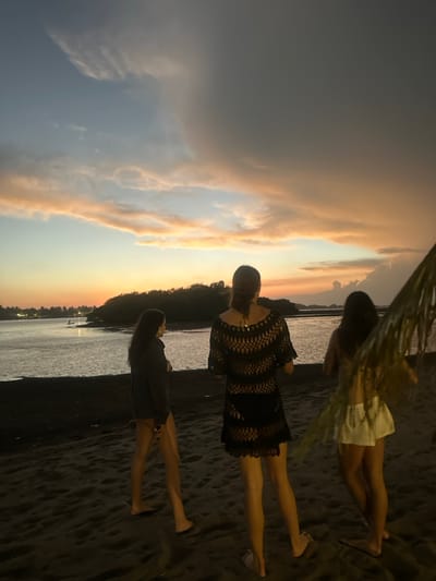 Three women stand on a dark sand beach under a vibrant sunset sky. The sky transitions from blue to orange with soft clouds. The woman in the middle wears a black crochet dress. Another woman wears a light-colored dress. The third woman's attire is dark. The beach slopes down to the water's edge. A green plant hangs at the top corner of the image.