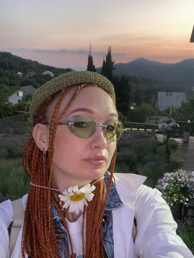 A close-up selfie shows a woman with bright orange braided hair and a green knit headband. She is wearing large, round, light green tinted sunglasses with silver metallic details around the lenses. Around her neck, she has a delicate beaded necklace adorned with a large white daisy with a yellow center. Her attire includes a white collared shirt layered over a denim vest and a white t-shirt. She is also wearing a long, silver chain earring in her left ear. The background reveals a scenic view of the city of Verkhneye Uchdere, Russia, with rolling green hills, scattered houses with glowing windows, and tall, dark trees. The sky displays a soft gradient of pink and orange hues, indicative of sunset. In the foreground, there are clusters of purple and white flowers, and a portion of a wooden deck can be seen.