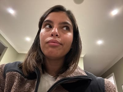 The image depicts an indoor scene focused on a young adult female. She has dark brown hair, brown eyes, and is looking upwards and to the right with a slightly pursed mouth, conveying a contemplative or pensive expression. She is wearing a light-colored (likely white) long-sleeved top underneath a brown, fuzzy sherpa-style vest or jacket with black fabric panels on the shoulders and collar. The background features a light-colored ceiling with multiple illuminated circular recessed lights. White crown molding runs along the top of a muted green or olive-colored wall. A partially visible doorway or opening to another room is present in the background to the right. The environment suggests a residential or office interior. No specific identifying markers for the city of Princeton, United States are visible in the image.